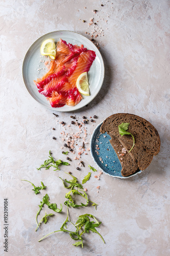 Sliced beetroot marinated salmon for sandwiches with sliced rye bread, pink salt, pepper, greens and lemon served in blue plates over grey texture background. Top view, space photo