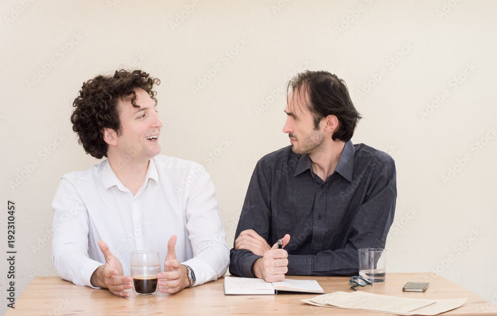 Two young Caucasian colleagues discussing at the table in the office with light background. Two young Caucasian colleagues discussing at the table in the office with light background.