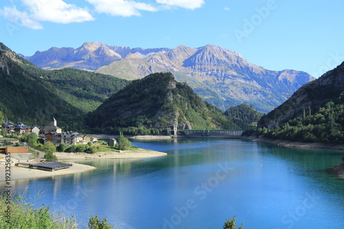 Pyrenees landscape at sunset. It shows a swamp, the village and a beach in the foreground; in the background the high mountains under a blue sky.
