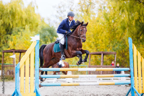 Young rider man on bay horse jumping over hurdle on show jumping competition