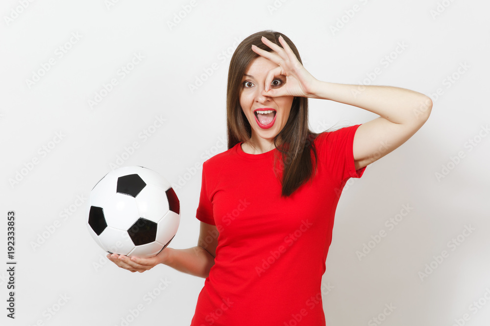 Pretty European young woman, football fan or player in red uniform holding classic soccer ball, fingers near eye isolated on white background. Sport, play football, health, healthy lifestyle concept.