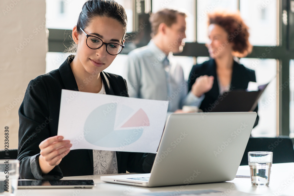 Hard-working young woman analyzing printed and electronic business information while sitting at desk in the office Hard-working young woman analyzing printed and electronic business information while sitting at desk in the office