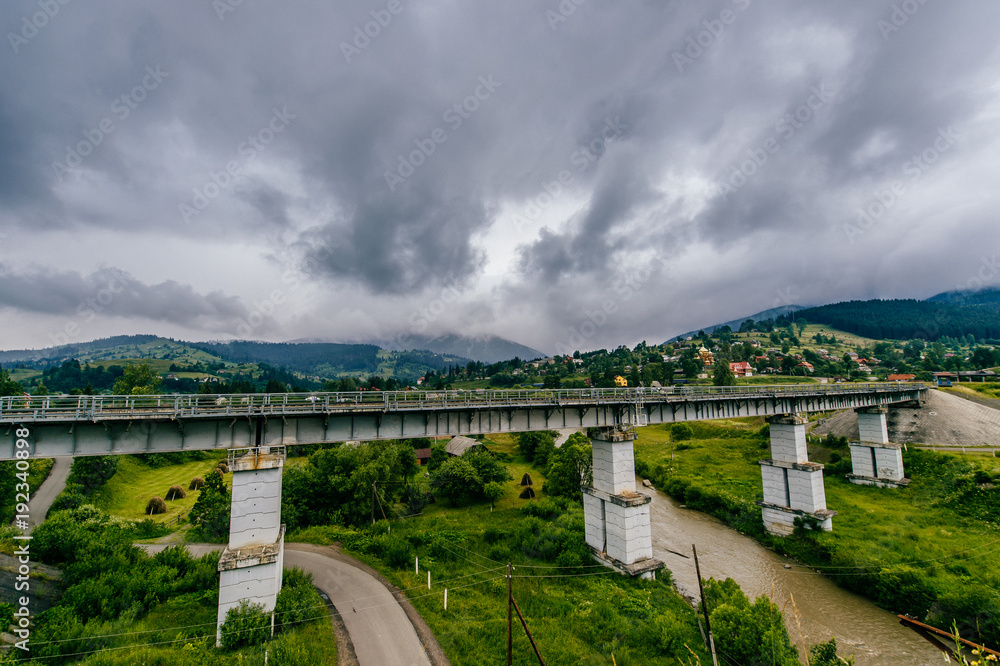Picturesque fairy tale landscape with a view at railway bridge and mountains covered with stormy rainy clouds. Fantasy colorful scenic route Hills and river. Tourist destination in Carpathians.