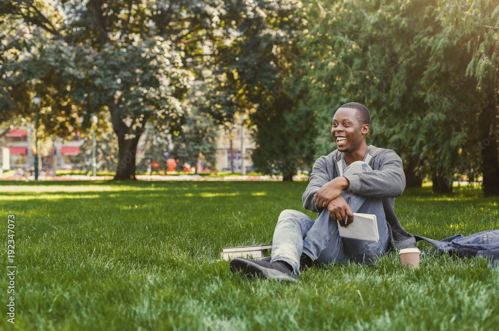 Happy african-american studen sitting with notebook on grass Happy african-american studen sitting with notebook on grass