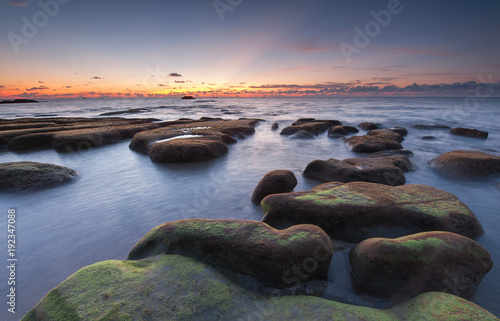 scenic view of sunset seascape with rocks covered by green moss on the ground. © udoikel09