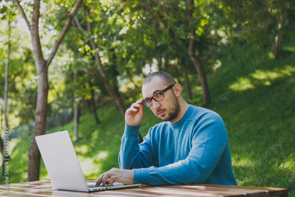 Young successful smart man businessman or student in casual blue shirt glasses sitting at table with mobile phone in city park using laptop working outdoors on green nature. Mobile Office concept. Young successful smart man businessman or student in casual blue shirt glasses sitting at table with mobile phone in city park using laptop working outdoors on green nature. Mobile Office concept.