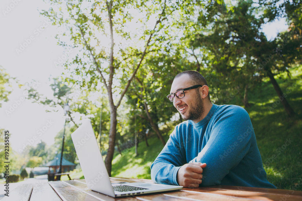 Young successful smart man businessman or student in casual blue shirt glasses sitting at table with mobile phone in city park using laptop working outdoors on green nature. Mobile Office concept. Young successful smart man businessman or student in casual blue shirt glasses sitting at table with mobile phone in city park using laptop working outdoors on green nature. Mobile Office concept.