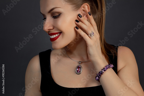 красивая молодая женщина с красной губной помадой и украшением на шее держит пальцы рук у лица, а сама она смотрит в сторону 