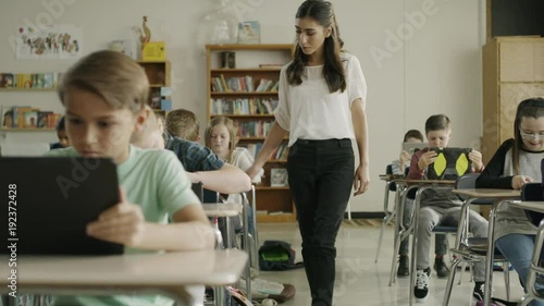 Panning shot of teacher assisting children in classroom reading digital tablets / Provo, Utah, United States