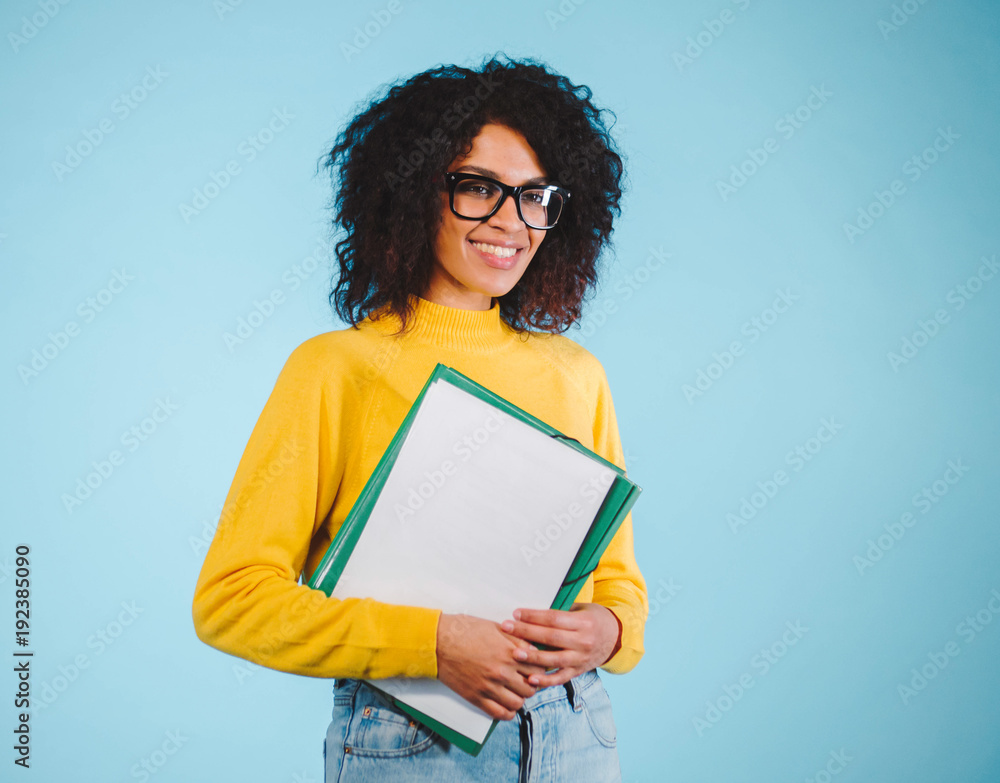 Obraz premium Portrait smiling young african woman in glasses holding folder over blue background. Education concept.