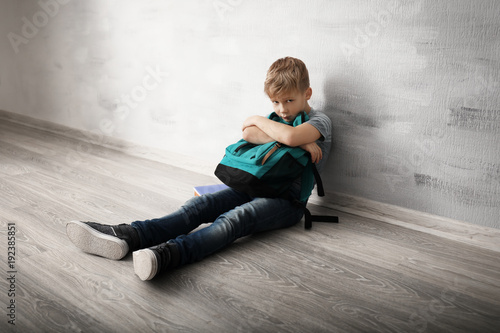 Upset little boy with backpack sitting on floor indoors. Bullying in school photo