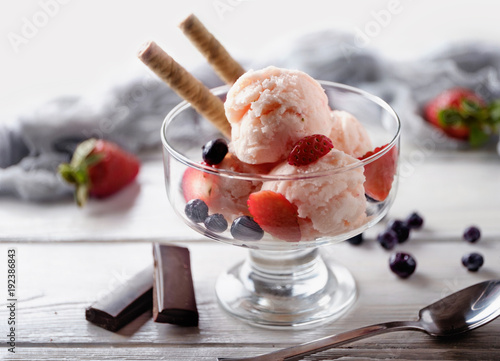 Several scoops of strawberry ice cream with wafer, strawberry and blue berry in glass against white wooden table. Summer background.