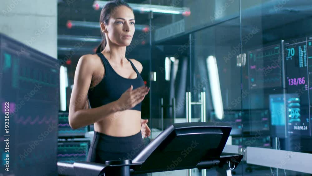 In Scientific Sports Laboratory Beautiful Woman Athlete Runs on a Treadmill with Electrodes Attached to Her Body, Monitors Show EKG Data on Display. Slow Motion. Out of Focus. In Scientific Sports Laboratory Beautiful Woman Athlete Runs on a Treadmill with Electrodes Attached to Her Body, Monitors Show EKG Data on Display. Slow Motion. Out of Focus.