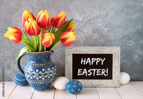 Red-yellow tulips in blue ceramic pitcher with Easter eggs and a blackboard  text