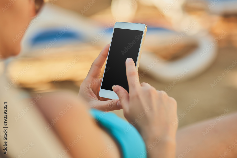 Close up view of a young attractive woman on holiday laying down on a white chaise-longue, holding and using a smartphone.