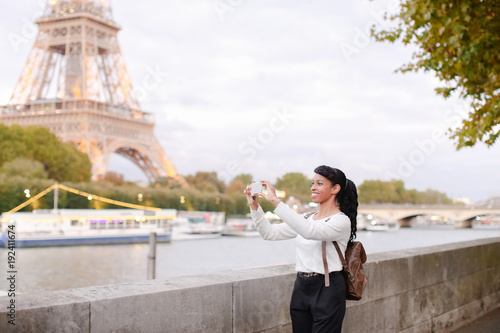 Student walking along embankment not far from Eiffel Tower and making photos of landmarks using smartphone. Young woman recently moved to Paris to live and study. Smiling black-haired female with