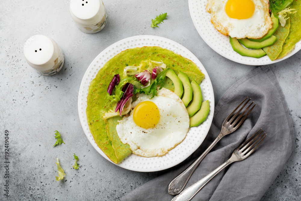 Spinach green crepes (pancakes) with fried egg, avocado and leaves of mix of salad on ceramic plate on gray concrete background. Сoncept of healthy breakfast. Selective focus. Top view. Copt space. Spinach green crepes (pancakes) with fried egg, avocado and leaves of mix of salad on ceramic plate on gray concrete background. Сoncept of healthy breakfast. Selective focus. Top view. Copt space.