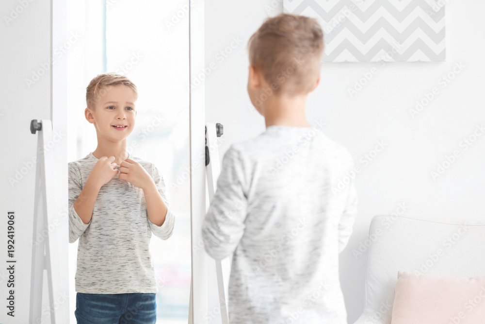 Cute little boy looking at himself in mirror indoors