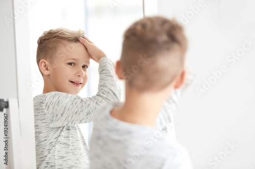 Cute little boy looking at himself in mirror indoors