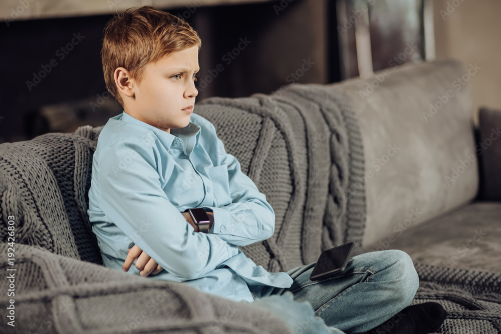 Down in dumps. Cute pre-teen boy sitting cross-legged on the sofa and folding his arms across his chest while looking disappointed, having lost a game Down in dumps. Cute pre-teen boy sitting cross-legged on the sofa and folding his arms across his chest while looking disappointed, having lost a game