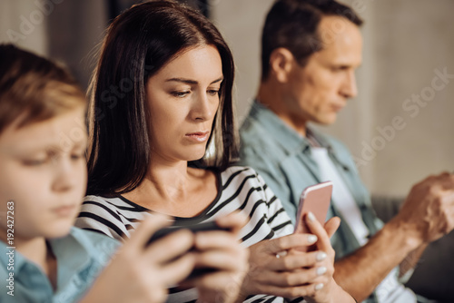 Totally concentrated. Pleasant young woman using her phone and being focused on it while sitting between her husband and son playing games