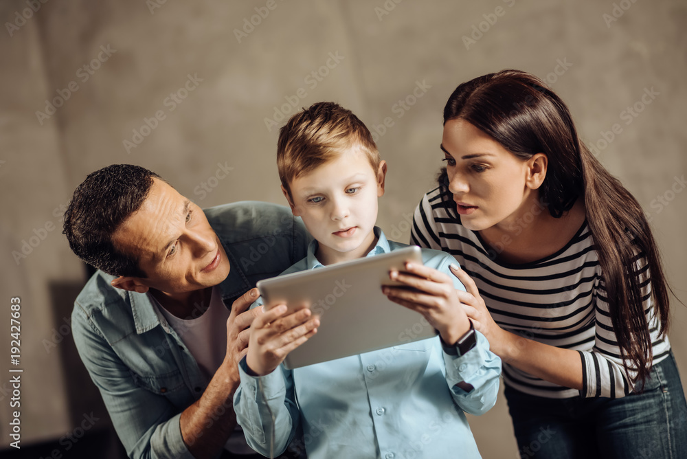 Serious concern. Loving young parents standing next to their pre-teen son and gently persuading him to stop playing on tablet while the boy taking no heed of them