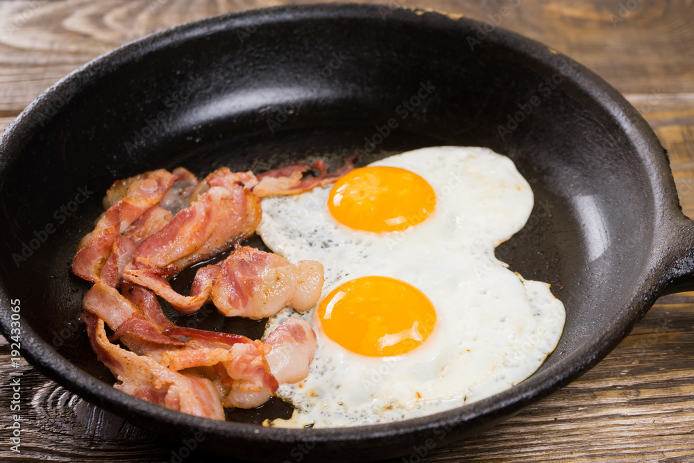 Bacon and Egg. Salted egg and sprinkled with black pepper. English breakfast. Grilled bacon, two eggs on pan on wooden table. Bacon and Egg. Salted egg and sprinkled with black pepper. English breakfast. Grilled bacon, two eggs on pan on wooden table.