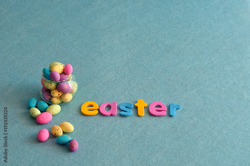 A small glass container filled with colorful speckled eggs and the word easter on a blue background A small glass container filled with colorful speckled eggs and the word easter on a blue background