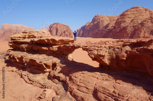 Young man standing at natural rock bridge and panoramic view of Jordanian desert Wadi Rum