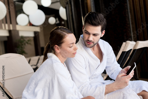 young couple in bathrobes using smartphone together in spa center