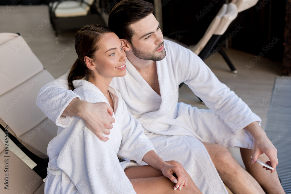 young couple in bathrobes embracing and looking away in spa center