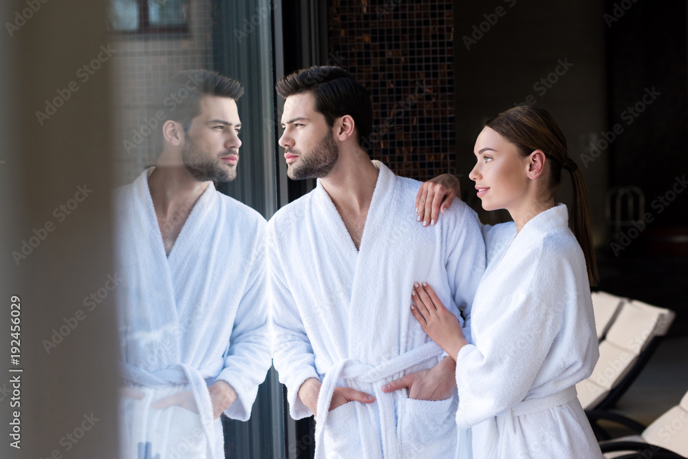 Obraz premium young couple in bathrobes standing together and looking away in spa center