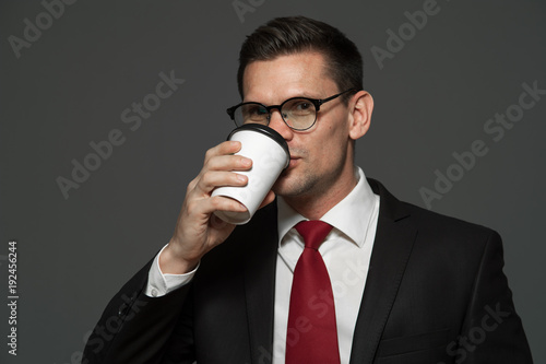 Portrait of young male manager in formal attire and glasses is drinking coffee on gray background