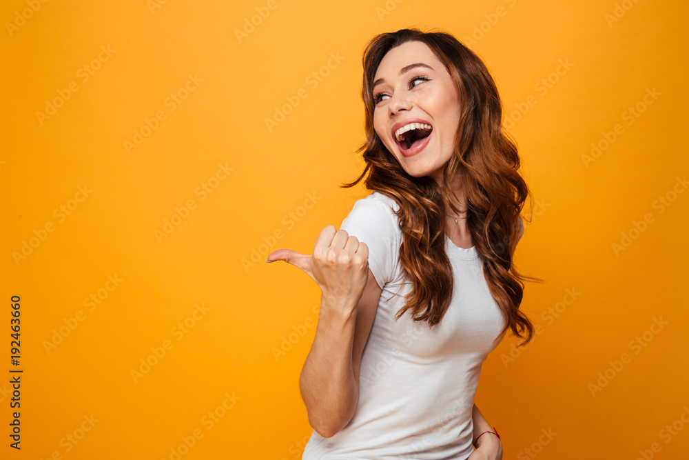 Laughing brunette woman in t-shirt looking and pointing away - obrazy, fototapety, plakaty 
