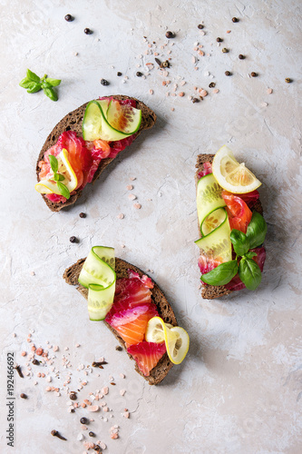 Sliced beetroot marinated salmon sandwiches with rye bread, cucumber, basil and lemon over grey texture background. Top view, space photo