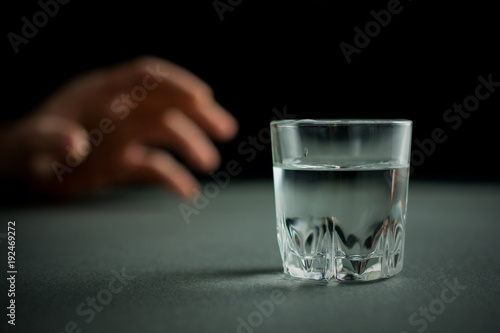 hand reaches for a glass of vodka or alcohol drink, alcoholism and alcohol abuse concept, defocused,.selective focus, close up, gray table, dark background
