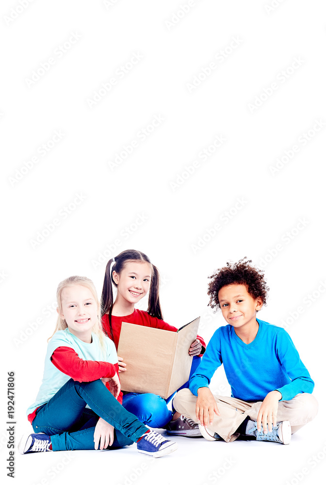 Three multi-ethnic elementary students sitting on floor and reading a book together Three multi-ethnic elementary students sitting on floor and reading a book together