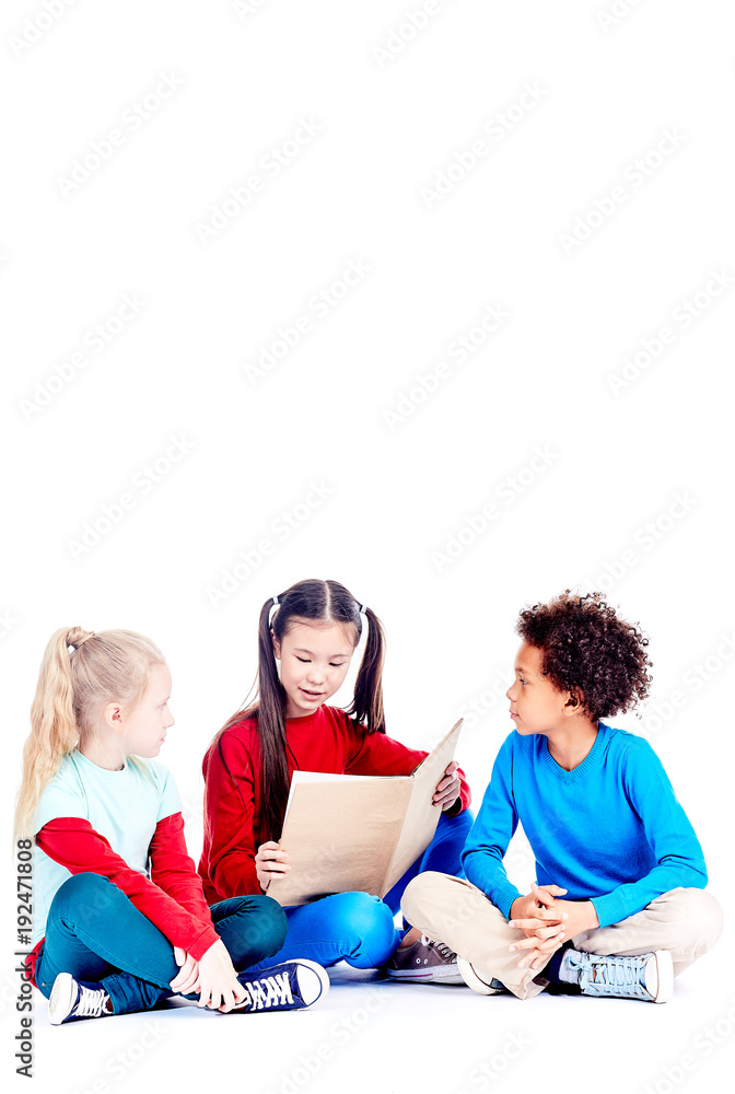 Three multi-ethnic elementary students sitting on floor and reading a book together Three multi-ethnic elementary students sitting on floor and reading a book together