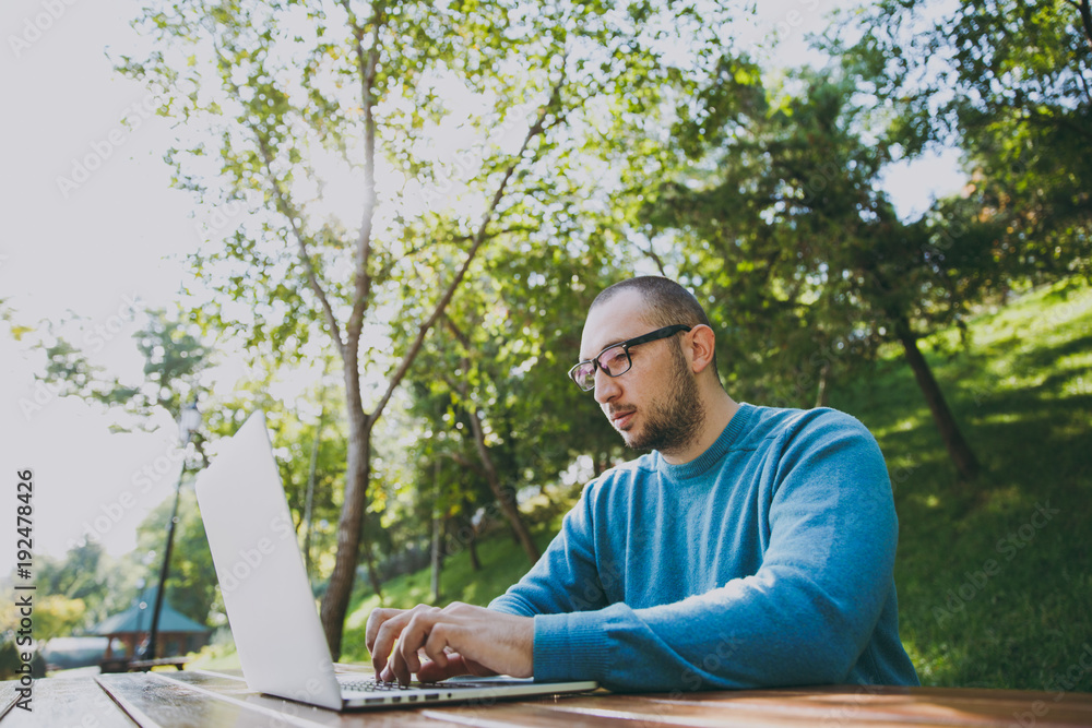 Young successful smart man businessman or student in casual blue shirt glasses sitting at table with mobile phone in city park using laptop working outdoors on green nature. Mobile Office concept. Young successful smart man businessman or student in casual blue shirt glasses sitting at table with mobile phone in city park using laptop working outdoors on green nature. Mobile Office concept.