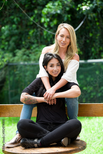A pair of proud lesbian in outdoors sitting on a wooden table, blonde woman is hugging a brunette woman, in a garden background photo