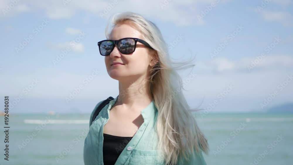 Young happy caucasian woman with long blonde hair in sunglasses and green shirt standing and smiling near palm tree on a blue sea background. Travel concept Young happy caucasian woman with long blonde hair in sunglasses and green shirt standing and smiling near palm tree on a blue sea background. Travel concept