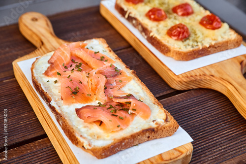 A set of sandwiches with melted cheese and sun dried tomatoes and baked cheese and salmon on a light cutting board and a brown wooden table. Photographed with natural light.
