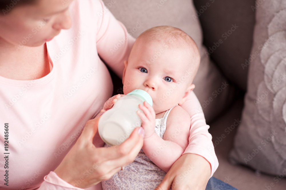 Mother feeding baby in room. Infant holding drinking milk from bottle closeup. Looking at camera. Healthy nutrition. Mother feeding baby in room. Infant holding drinking milk from bottle closeup. Looking at camera. Healthy nutrition.