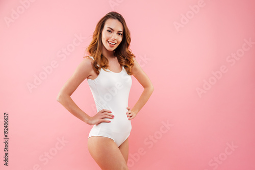 Beautiful slim girl 20s with brown hair wearing one-piece swimsuit smiling and posing on camera keeping hands on waist isolated over pink background