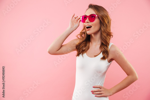 Sexual magnificent woman with brown hair in swimsuit and sunglasses smiling and looking aside isolated over pink background