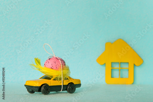 A yellow car with an egg on the roof and a house on a blue background. Easter concepts. Holiday for Easter. Travel to Easter holidays.