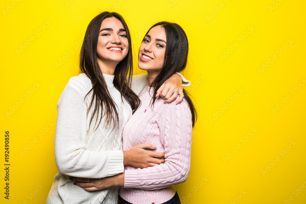 Portrait of two happy girls dressed in sweaters hugging isolated over yellow background Portrait of two happy girls dressed in sweaters hugging isolated over yellow background