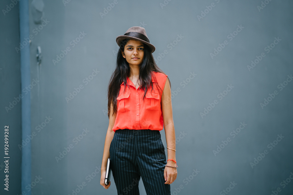 Portrait of a fashionable, young and attractive Indian woman standing outdoors against a blue grey background and looking up. She is dressed fashionably with a hat, holding a notebook. She looks cheer Portrait of a fashionable, young and attractive Indian woman standing outdoors against a blue grey background and looking up. She is dressed fashionably with a hat, holding a notebook. She looks cheer