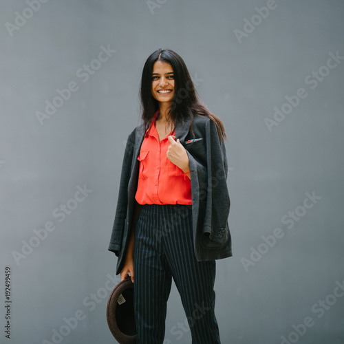 A fashionable and attractive young Indian Asian a woman poses against a plain grey background and smiling. She is wearing a jacket with a paisley pocket square and holding a fedora hat. 