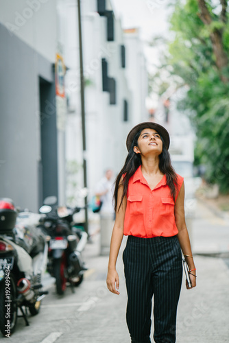 Portrait of a young Indian Asian model woman walking along a street in the day and smiling. She is dressed fashionably and stylishly in smart casual clothes and a fedora hat in the summer sun.